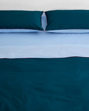 A neatly made bed features two dark teal pillows and the Bed Threads Aegean, Sky & Bubblegum Piped 100% Organic Cotton Duvet Cover, accented by light blue sheets and piping. The background wall is white. - view