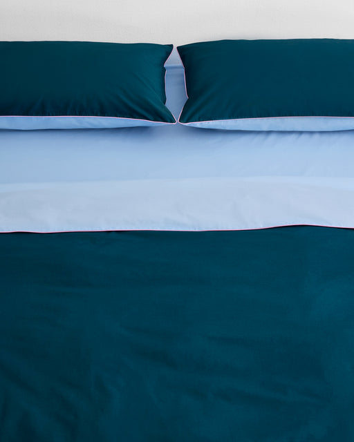 A neatly made bed features two dark teal pillows and the Bed Threads Aegean, Sky & Bubblegum Piped 100% Organic Cotton Duvet Cover, accented by light blue sheets and piping. The background wall is white.