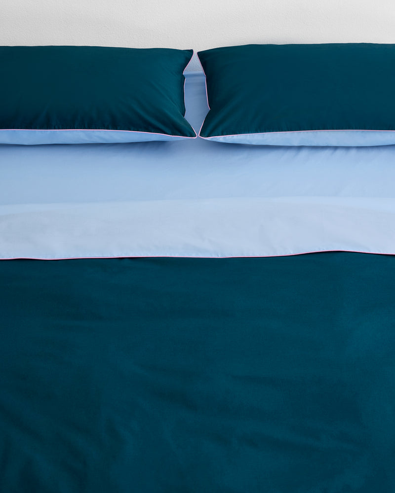 A neatly made bed features two dark teal pillows and the Bed Threads Aegean, Sky & Bubblegum Piped 100% Organic Cotton Duvet Cover, accented by light blue sheets and piping. The background wall is white.