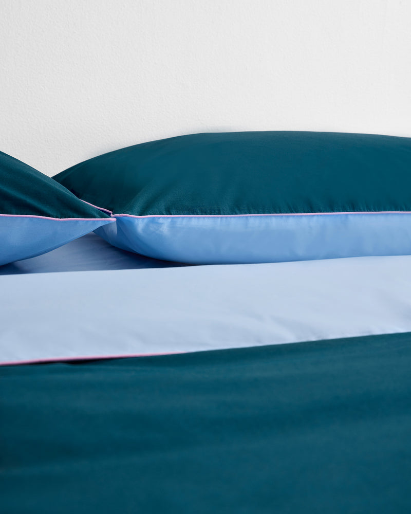 A bed with Bed Threads' Aegean, Sky & Bubblegum Piped 100% Organic Cotton Pillowcases (GOTS-certified) showcases teal and light blue hues, pink piping accents, and rests against a plain white wall.