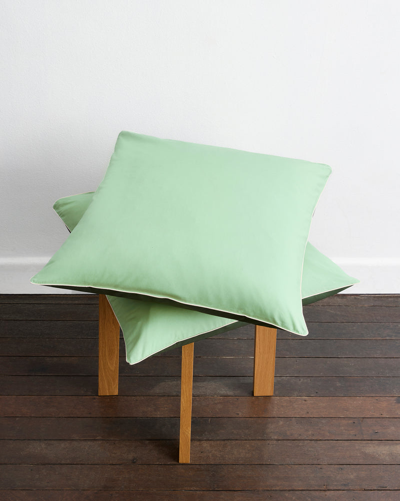 Two Moss, Apple & Vanilla Piped 100% Organic Cotton European Pillowcases by Bed Threads are stacked on a wooden stool, placed on a dark floor against a white wall.