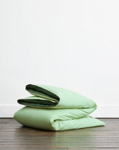 The Moss, Apple & Vanilla Piped 100% Organic Cotton Duvet Cover Set by Bed Threads is neatly folded on a wooden floor against a plain white wall. - view