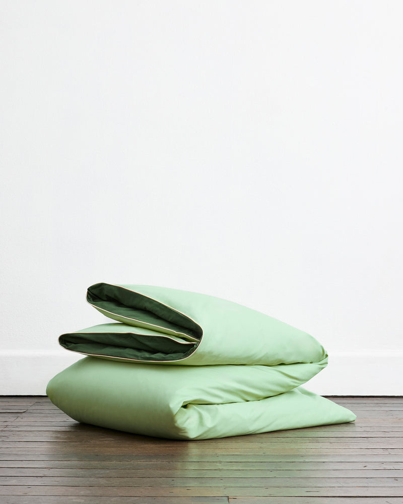The Moss, Apple & Vanilla Piped 100% Organic Cotton Duvet Cover Set by Bed Threads is neatly folded on a wooden floor against a plain white wall.