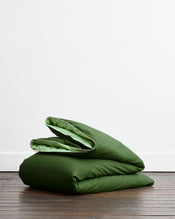 A modern bedroom with a green bed, wood-paneled walls, large windows, brown curtains and rug, minimalist bedside tables with lamps and glassware, and Bed Threads' Moss, Apple & Vanilla Piped 100% Organic Cotton European Pillowcases (Set of Two). - view
