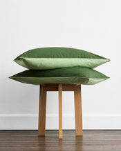 A neatly made bed features Bed Threads' Moss, Apple & Vanilla Piped 100% Organic Cotton European Pillowcases (Set of Two) in light green, paired with dark green shams and a runner, all set against a wooden headboard. - view