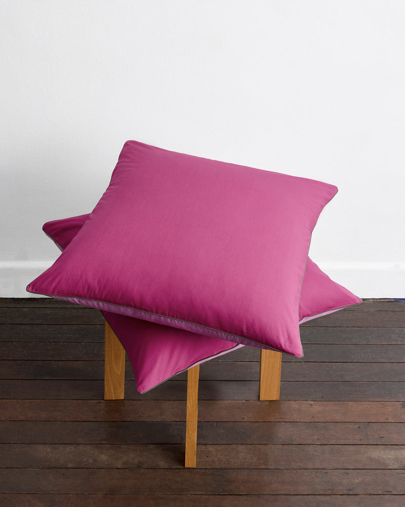A neatly made bed with Berry, Bubblegum & Moss Piped 100% Organic Cotton European Pillowcases (Set of Two) by Bed Threads sits against a wooden headboard, sunlight partially illuminating the light pink and magenta bedding.