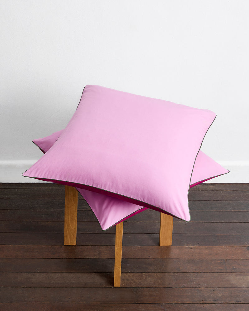 A set of Berry, Bubblegum & Moss Piped 100% Organic Cotton European Pillowcases by Bed Threads is stacked on a simple wooden stool placed on a dark floor against a plain white wall.