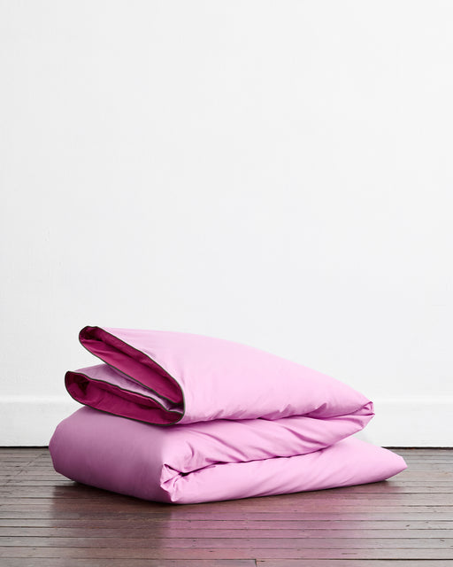 The Berry, Bubblegum & Moss Piped 100% Organic Cotton Bedding Bundle by Bed Threads, featuring a folded pink comforter with purple lining, rests on a wooden floor against a plain white wall.