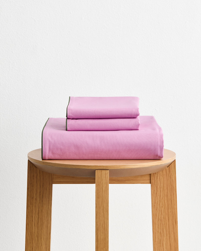The Berry, Bubblegum & Moss Piped 100% Organic Cotton Duvet Cover Set by Bed Threads, featuring pink GOTS-certified bedding with green trim, is neatly folded on a light wooden stool against a plain white background.