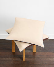 Two large pillows with Bed Threads’ Espresso, Vanilla & Sky Piped 100% Organic Cotton European Pillowcases are stacked askew on a four-legged wooden stool, set on a dark floor against a plain white wall. - view
