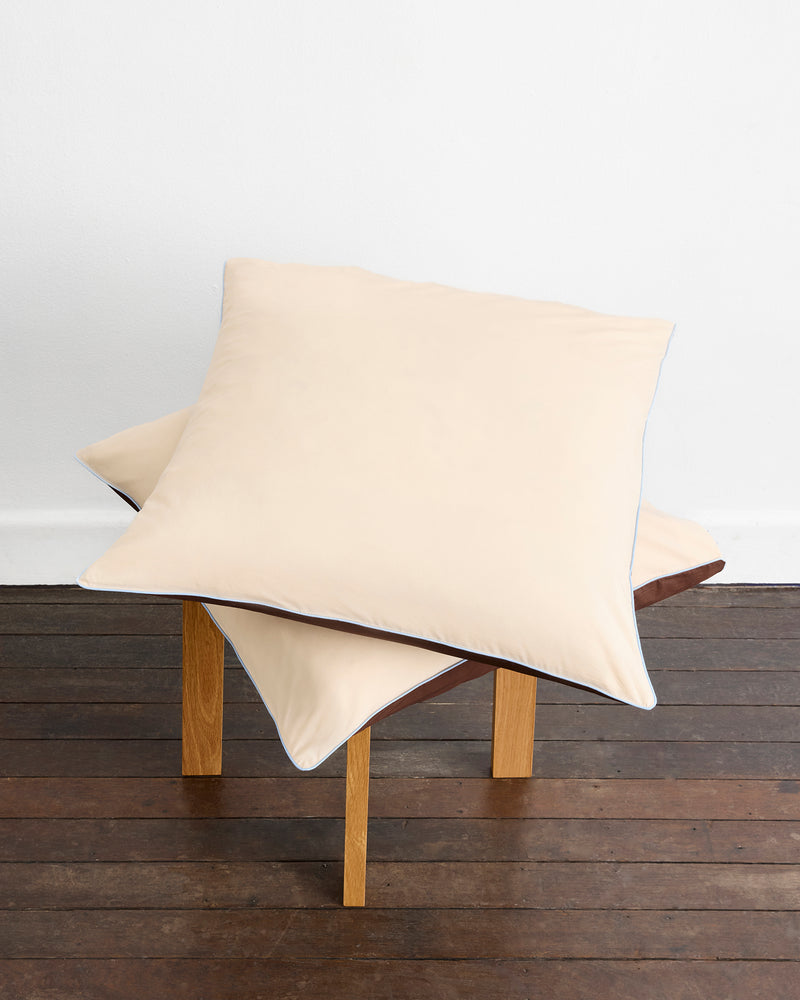 Two large pillows with Bed Threads’ Espresso, Vanilla & Sky Piped 100% Organic Cotton European Pillowcases are stacked askew on a four-legged wooden stool, set on a dark floor against a plain white wall.