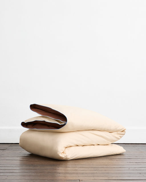 The Bed Threads Espresso, Vanilla & Sky Piped 100% Organic Cotton Duvet Cover is folded and cream-colored with brown piping, displayed on a wooden floor against a plain white wall.