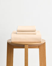 The Bed Threads Espresso, Vanilla & Sky Piped 100% Organic Cotton Duvet Cover Set, neatly folded, rests on a wooden stool against a plain white wall. - view