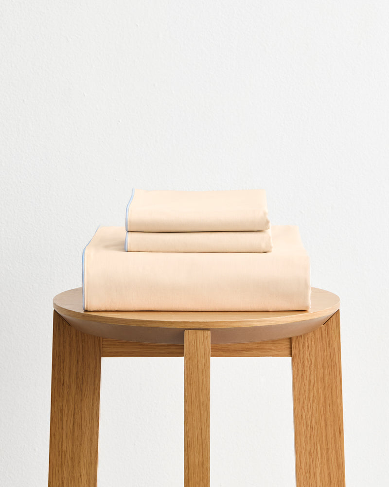 The Bed Threads Espresso, Vanilla & Sky Piped 100% Organic Cotton Duvet Cover Set, neatly folded, rests on a wooden stool against a plain white wall.