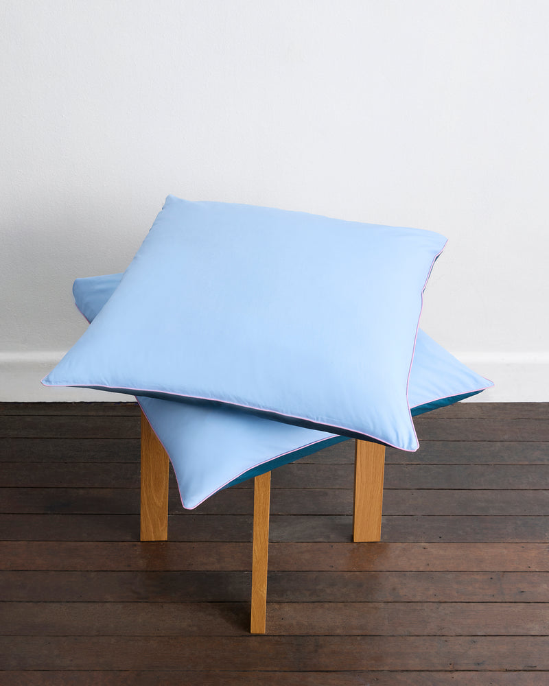 Two Aegean, Sky & Bubblegum Piped 100% Organic Cotton European Pillowcases by Bed Threads are stacked on a small wooden table with a dark wood floor and plain white wall in the background. Set includes two pillowcases.