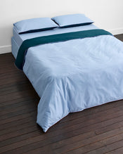 A neatly made bed features Bed Threads’ Aegean, Sky & Bubblegum Piped 100% Organic Cotton Duvet Cover atop light blue percale sheets and pillowcases, with a dark teal blanket on a dark wooden floor against a plain white wall. - view