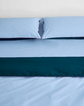 A neatly made bed with light blue percale sheets and pillows, topped with a dark teal runner and finished with the Bed Threads Aegean, Sky & Bubblegum Piped 100% Organic Cotton Duvet Cover, set against a plain white wall. - view