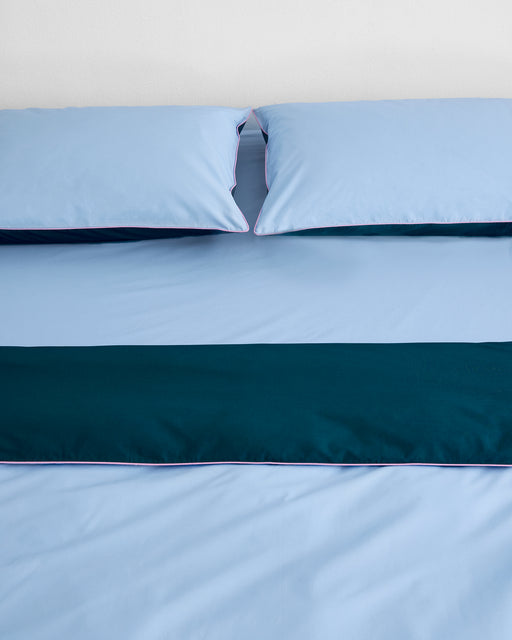 A neatly made bed with light blue percale sheets and pillows, topped with a dark teal runner and finished with the Bed Threads Aegean, Sky & Bubblegum Piped 100% Organic Cotton Duvet Cover, set against a plain white wall.