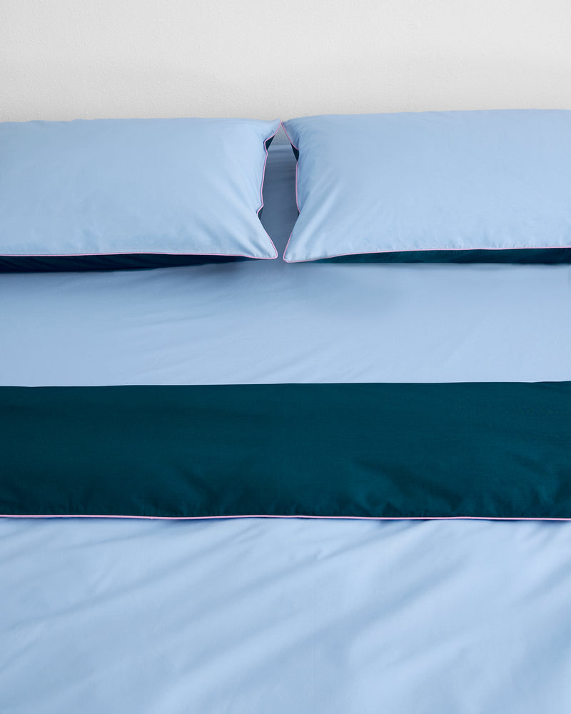 A neatly made bed with light blue percale sheets and pillows, topped with a dark teal runner and finished with the Bed Threads Aegean, Sky & Bubblegum Piped 100% Organic Cotton Duvet Cover, set against a plain white wall.