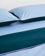 Close-up of Bed Threads’ Aegean, Sky & Bubblegum Piped 100% Organic Cotton Pillowcases (Set of Two) on a neatly made bed, featuring light blue and teal bedding with pink piping for a clean, modern look. - view