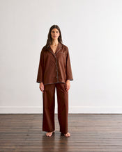 A person stands barefoot on a wooden floor, wearing the Bed Threads Espresso & Sky Piped Long Sleep Set—loose-fitting pants with an elastic waistband and side pockets—against a plain white wall. - view