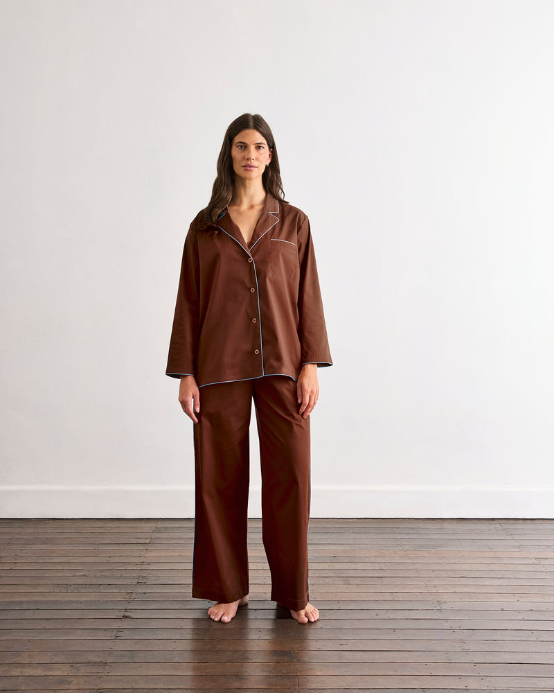 A person stands barefoot on a wooden floor, wearing the Bed Threads Espresso & Sky Piped Long Sleep Set—loose-fitting pants with an elastic waistband and side pockets—against a plain white wall.