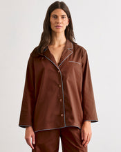 A woman faces away from the camera, wearing the Bed Threads Espresso & Sky Piped Long Sleeve Shirt—a brown cotton pyjama top with light blue piping—paired with matching wide-leg pants. - view