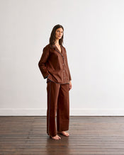 A woman with long brown hair stands barefoot on a wooden floor against a white wall, wearing Bed Threads' Espresso & Sky Piped Pants—a brown pajama set with white piping and relaxed, elastic-waist pants. - view