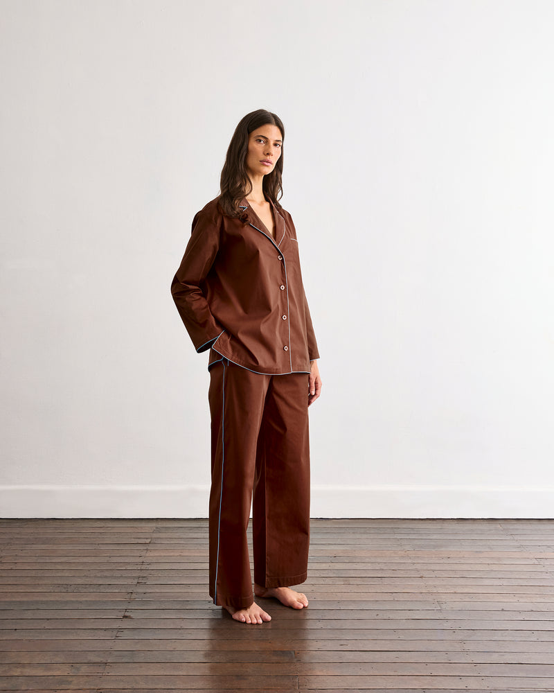 A woman with long brown hair stands barefoot on a wooden floor against a white wall, wearing Bed Threads' Espresso & Sky Piped Pants—a brown pajama set with white piping and relaxed, elastic-waist pants.
