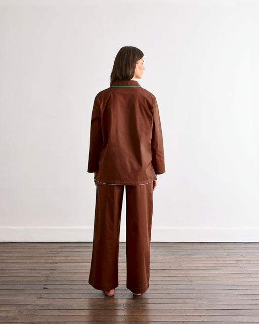 A woman stands barefoot on a wooden floor facing away, wearing the Bed Threads Espresso & Sky Piped Long Sleep Set—loose-fitting pants and matching long-sleeve top—against a plain white wall.
