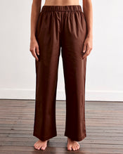 A woman with long brown hair stands barefoot on a wooden floor against a white wall, wearing Bed Threads' Espresso & Sky Piped Pants—a brown pajama set with white piping and relaxed, elastic-waist pants. - view
