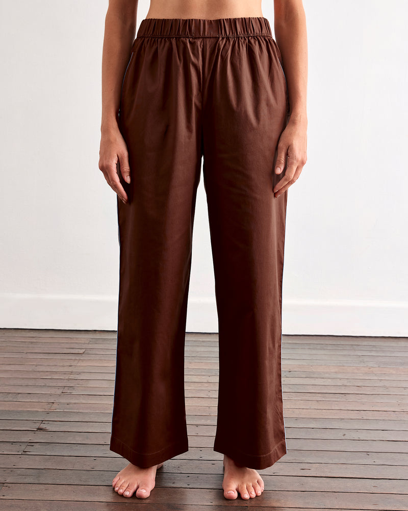 A woman with long brown hair stands barefoot on a wooden floor against a white wall, wearing Bed Threads' Espresso & Sky Piped Pants—a brown pajama set with white piping and relaxed, elastic-waist pants.