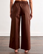 A person stands barefoot on wooden floorboards, seen from behind, wearing Bed Threads' Espresso & Sky Piped Pants with an elastic waist in breathable cotton. A plain white wall forms the background. - view