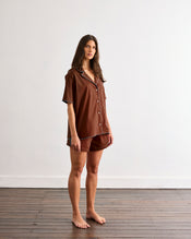 A woman with long brown hair stands barefoot on a wooden floor, wearing the Bed Threads Espresso & Sky Piped Short Sleeve Shirt—ideal for warmer nights—against a plain white wall. - view