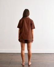 A woman with long brown hair stands barefoot on a wooden floor, facing a white wall. She is wearing the Bed Threads Espresso & Sky Piped Short Sleep Set, featuring a brown short sleeve shirt and matching piped shorts. - view
