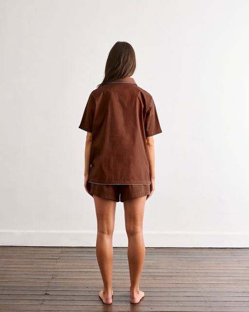 A woman with long brown hair stands barefoot on a wooden floor, facing a white wall. She is wearing the Bed Threads Espresso & Sky Piped Short Sleep Set, featuring a brown short sleeve shirt and matching piped shorts.