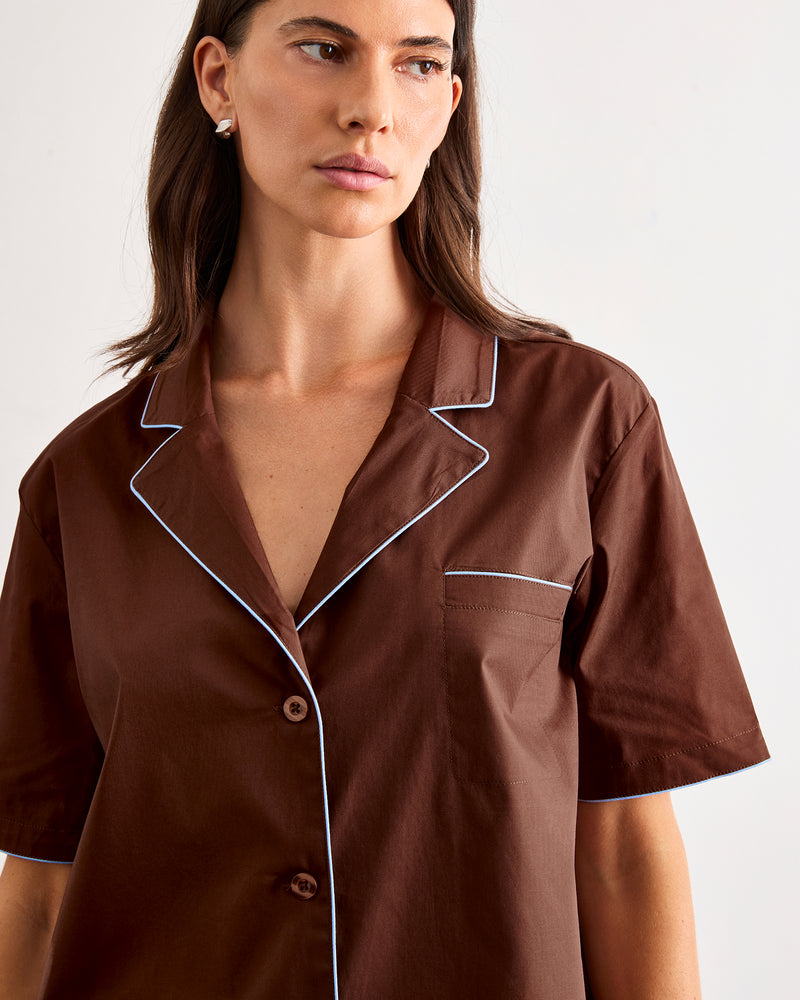 A woman with long brown hair wears the Bed Threads Espresso & Sky Piped Short Sleeve Shirt, a lightweight cotton pyjama top in brown with white piping along the collar, pocket, and sleeves—ideal for warmer nights.