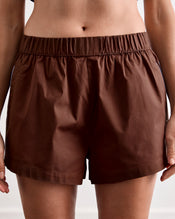 A woman stands barefoot on a wooden floor against a plain white wall, wearing Bed Threads’ Espresso & Sky Piped Shorts pajama set with white piping and breathable cotton fabric. - view
