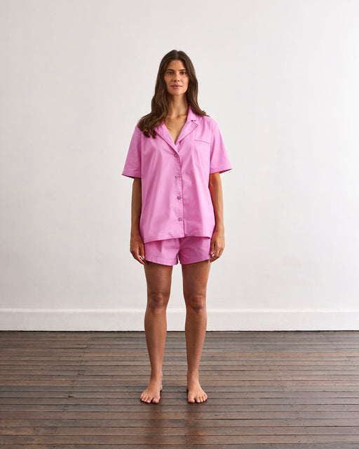 A person wearing the Bubblegum & Berry Piped Short Sleep Set by Bed Threads stands indoors; only their lower torso and upper legs in the elastic-waist shorts are visible.