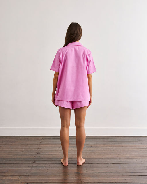 A person with long brown hair stands barefoot on a wooden floor, facing away from the camera, wearing the Bed Threads Bubblegum & Berry Piped Short Sleep Set against a plain white wall.