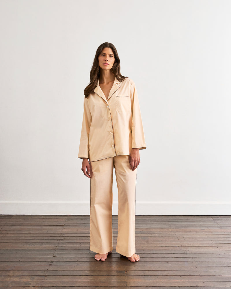 A person stands barefoot on a wooden floor, wearing loose, light beige pajama pants from the Vanilla & Espresso Piped Long Sleep Set by Bed Threads. The photo is cropped to show only the lower half of their body.