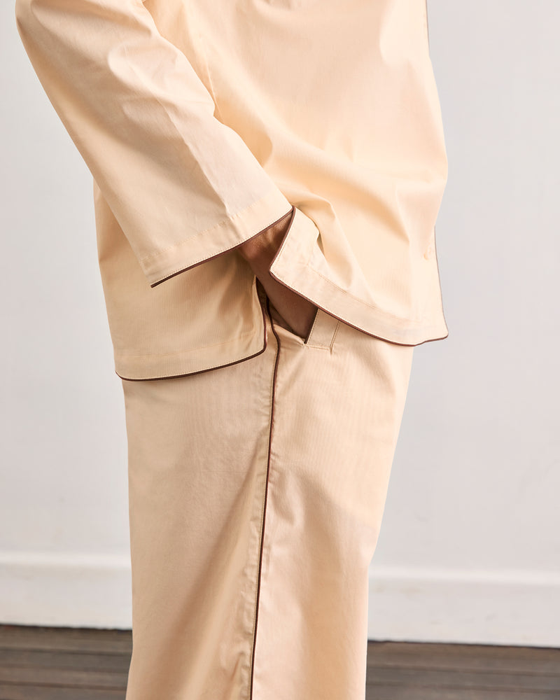 Person wearing Bed Threads Vanilla & Espresso Piped Pants, made from breathable cotton with brown piping and an elasticated waist, stands with one hand in their pocket against a plain white wall and wooden floor. Only torso and upper legs are visible.