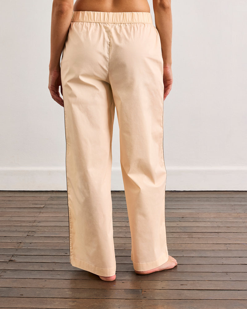 A person stands barefoot on a wooden floor, modeling Bed Threads’ Vanilla & Espresso Piped Pants—relaxed cotton trousers with a comfy elastic waist—shown from the back against a plain white wall.