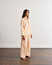 A woman stands barefoot on a wooden floor, wearing Bed Threads’ Vanilla & Espresso Piped Pants, crafted from breathable cotton with a relaxed fit and elasticated waist, set against a plain white wall. - view