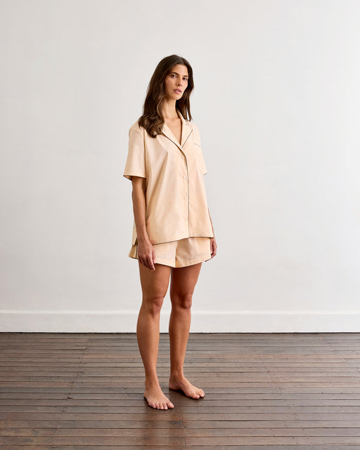 A woman stands barefoot on a wooden floor, wearing the Bed Threads Vanilla & Espresso Piped Short Sleeve Shirt and matching shorts, made of lightweight cotton. She poses against a plain white wall, looking slightly to the side.