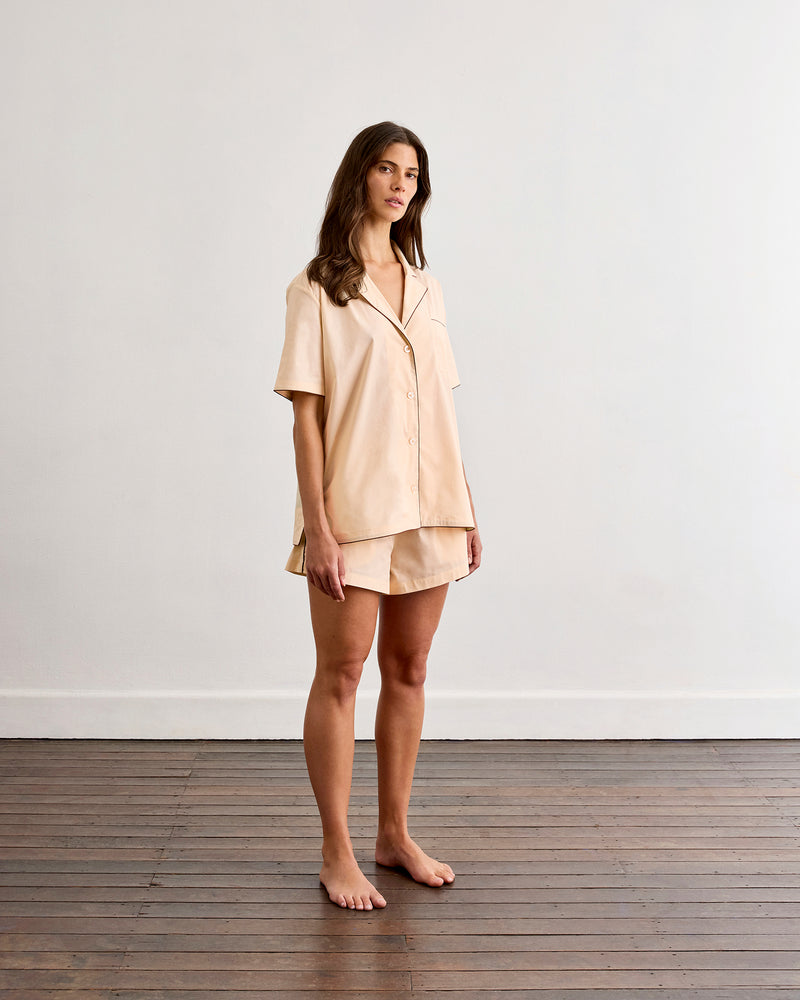 A woman stands barefoot on a wooden floor, wearing the Bed Threads Vanilla & Espresso Piped Short Sleeve Shirt and matching shorts, made of lightweight cotton. She poses against a plain white wall, looking slightly to the side.