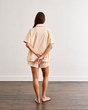 A woman stands barefoot on a wooden floor facing a white wall, hands behind her back. She wears the Vanilla & Espresso Piped Short Sleep Set by Bed Threads—a lightweight cotton set with a short sleeve shirt and matching piped shorts in beige. - view