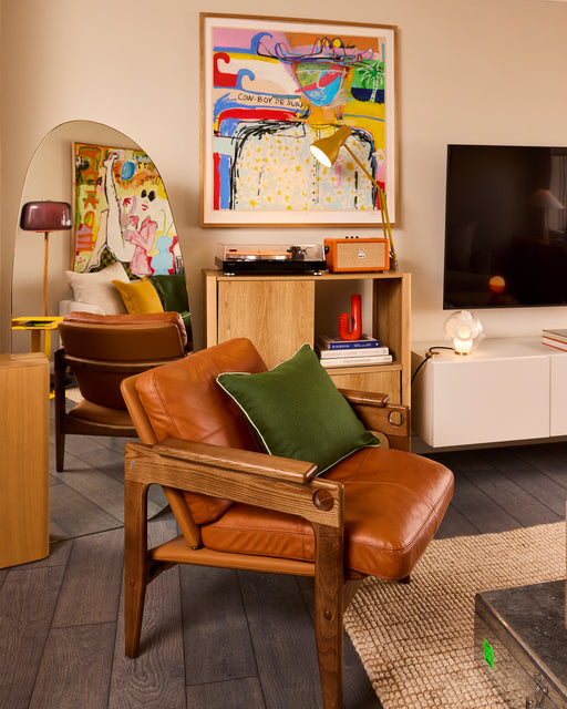 A cozy living room with a brown leather armchair, Bed Threads’ Olive & Sage 100% French Flax Linen Piped Cushion Cover, wooden shelves, modern decor, colorful abstract art, a round mirror, a TV, and warm ambient lighting.