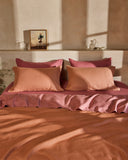 A neatly made bed with peach and mauve linens features Bed Threads Pink Clay 100% French Flax Linen Pillowcases. Sunlight casts soft shadows, and a shelf above displays a framed flower painting, a glass, and a white ceramic pitcher.