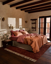 A bed dressed in the Bed Threads Hazelnut & Pink Clay 100% French Flax Linen Bedding Bundle sits against a beige wall, with a shelf above holding decor like a framed painting, glass, white vase, and ceramic pitcher. Shadows fall across the scene. - view